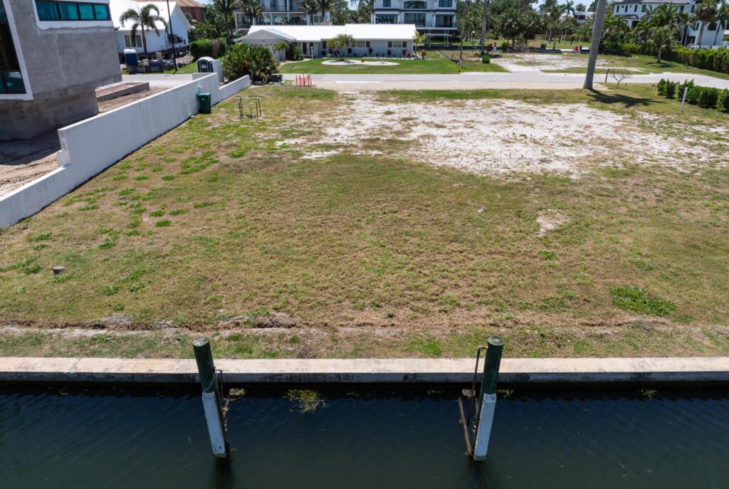 Empty waterfront lot with grass and sandy patches next to a canal.