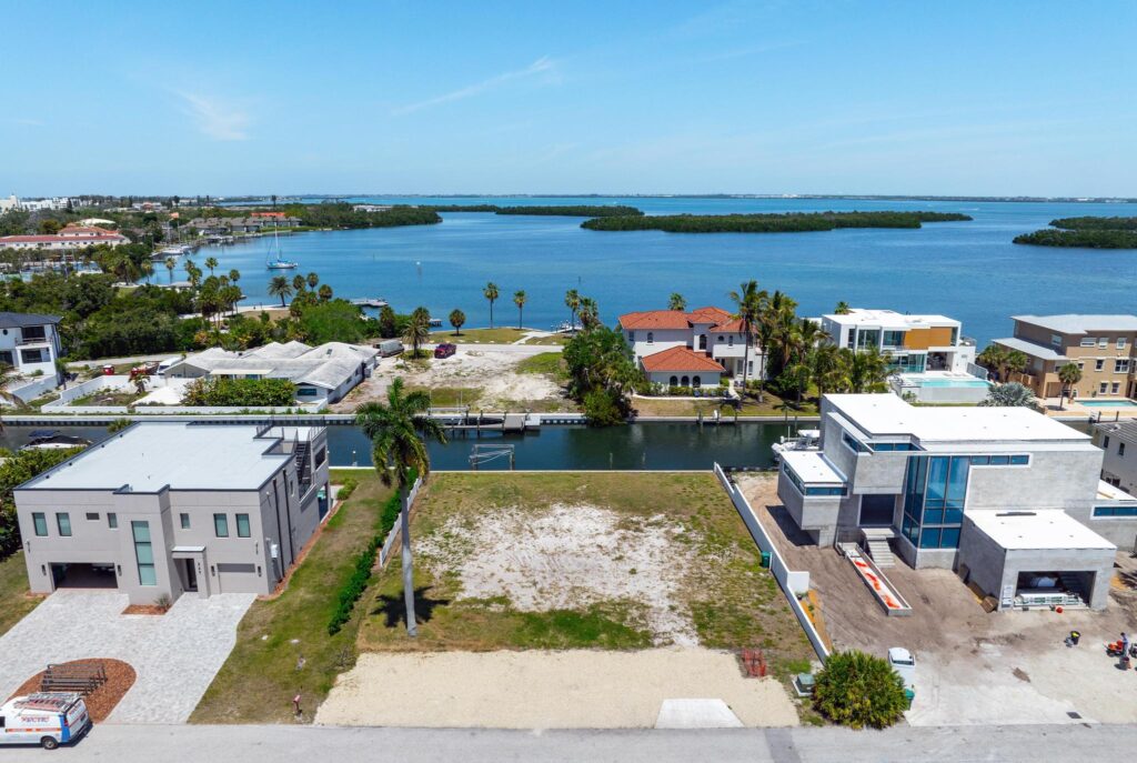 Empty waterfront lot with modern houses and clear blue sky.