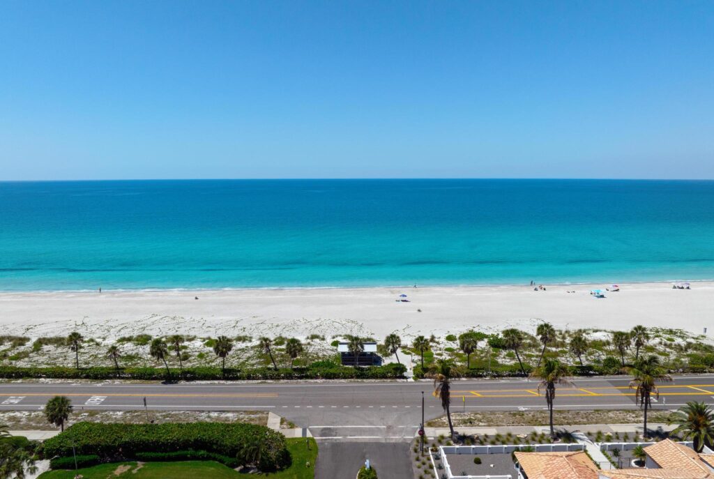 Clear blue ocean under a bright sky with a sandy beach and road in the foreground.