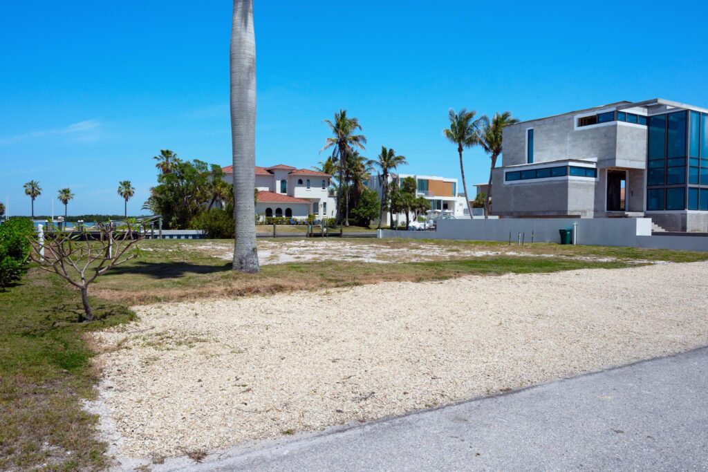 Empty lot with gravel near coastal homes and palm trees.